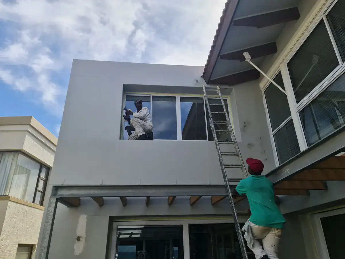 Painters preparing upper exterior walls around windows using ladders at a private Plattekloof home