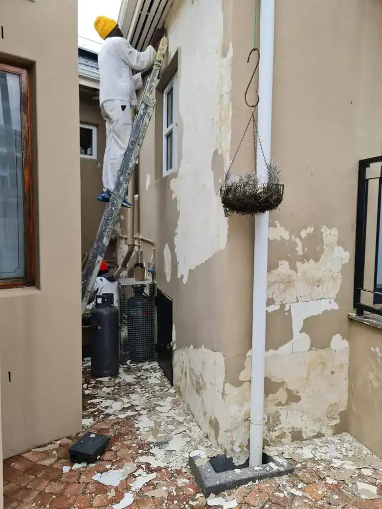 Painter removing loose peeling paint from damp-affected exterior walls at a heritage home in Tamboerskloof