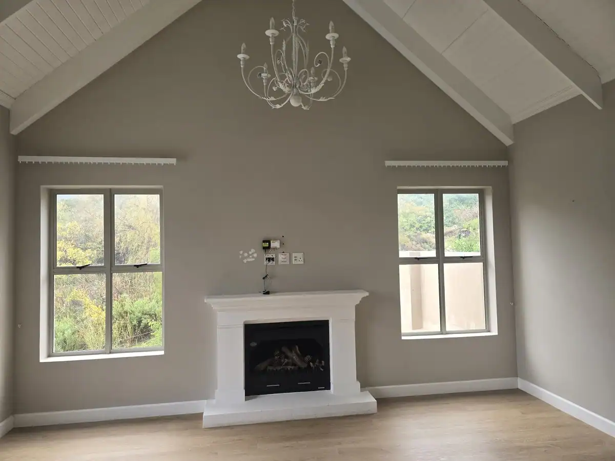 Double-volume living room with painted walls, white exposed roof trusses and fireplace after interior painting at L’Ermitage Estate in Franschhoek